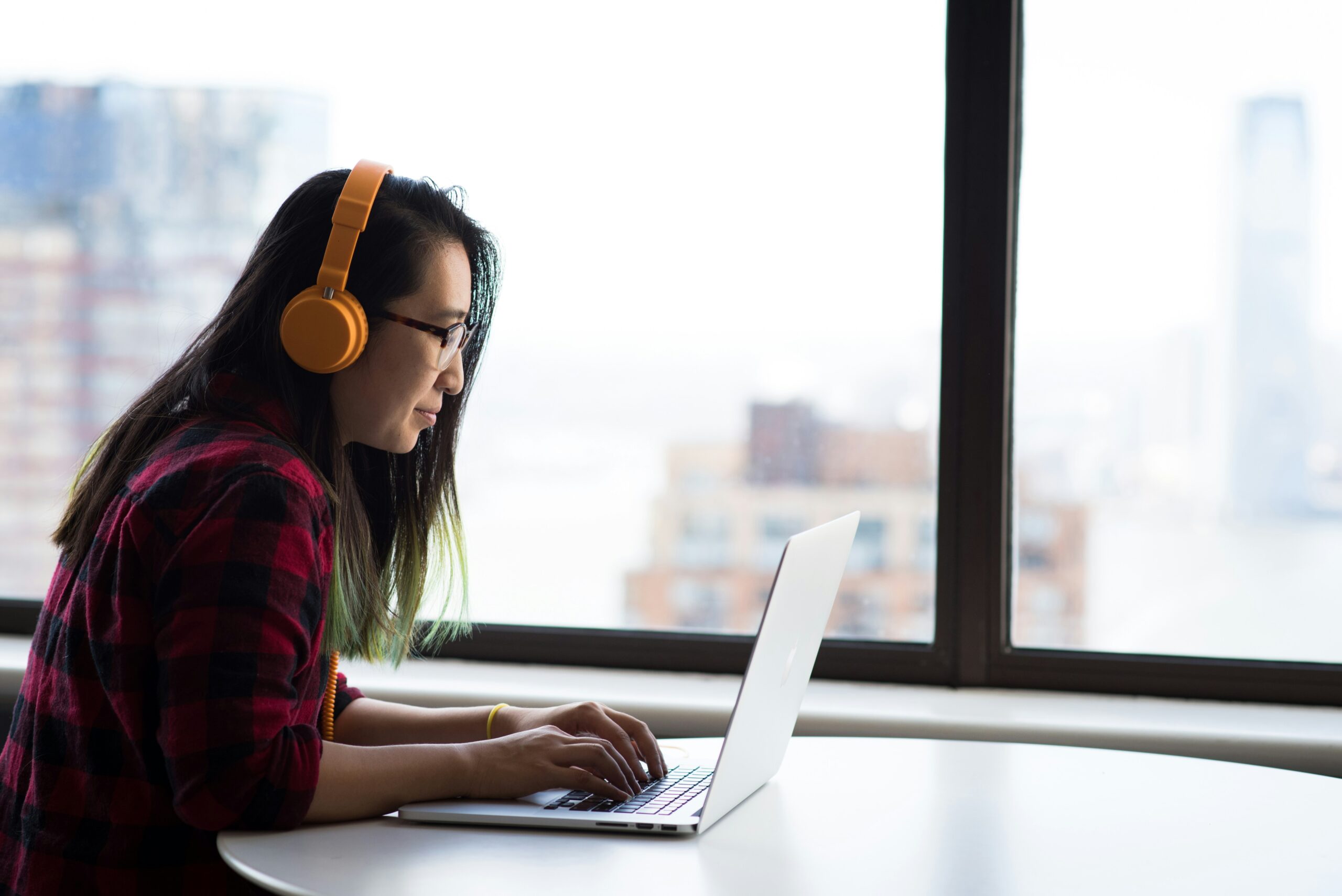 A focused woman wearing orange headphones and a red plaid shirt works on her laptop near a window, representing the benefits of custom development over templates for tailored and scalable digital solutions.