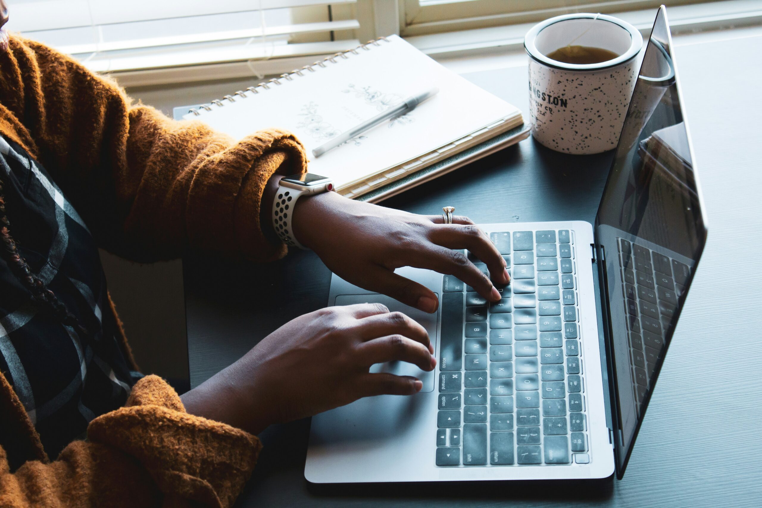 A person typing on a laptop with a notebook and coffee nearby, illustrating the flexibility, efficiency, and accelerated content creation and publishing capabilities of CMS for website management.