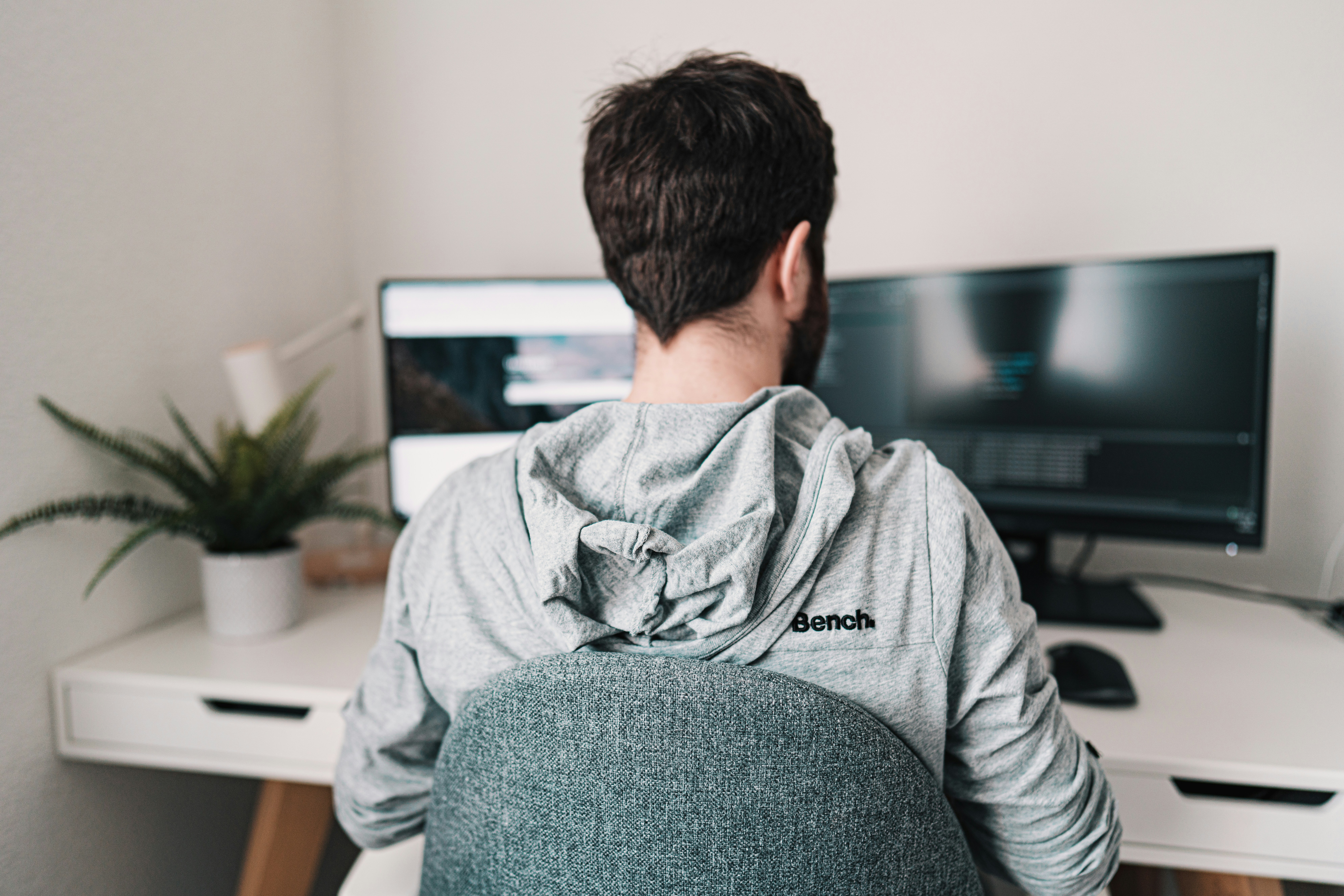 A person in a gray hoodie sitting at a desk with dual monitors displaying code, representing the process of analyzing remote front-end development services.