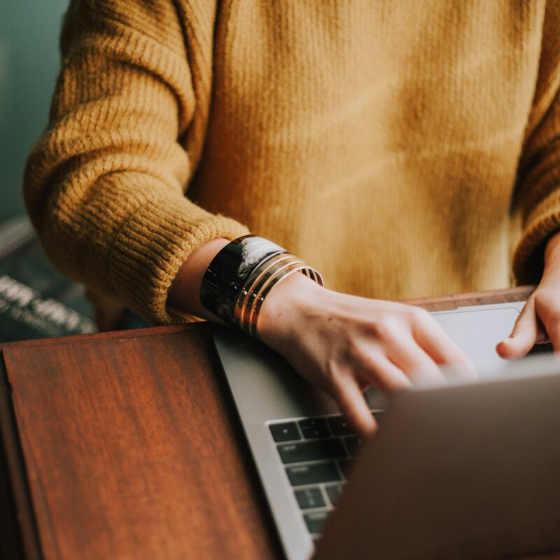 Person in a mustard sweater typing on a laptop at a wooden desk, symbolizing focused e-commerce keyword research to improve online search rankings.