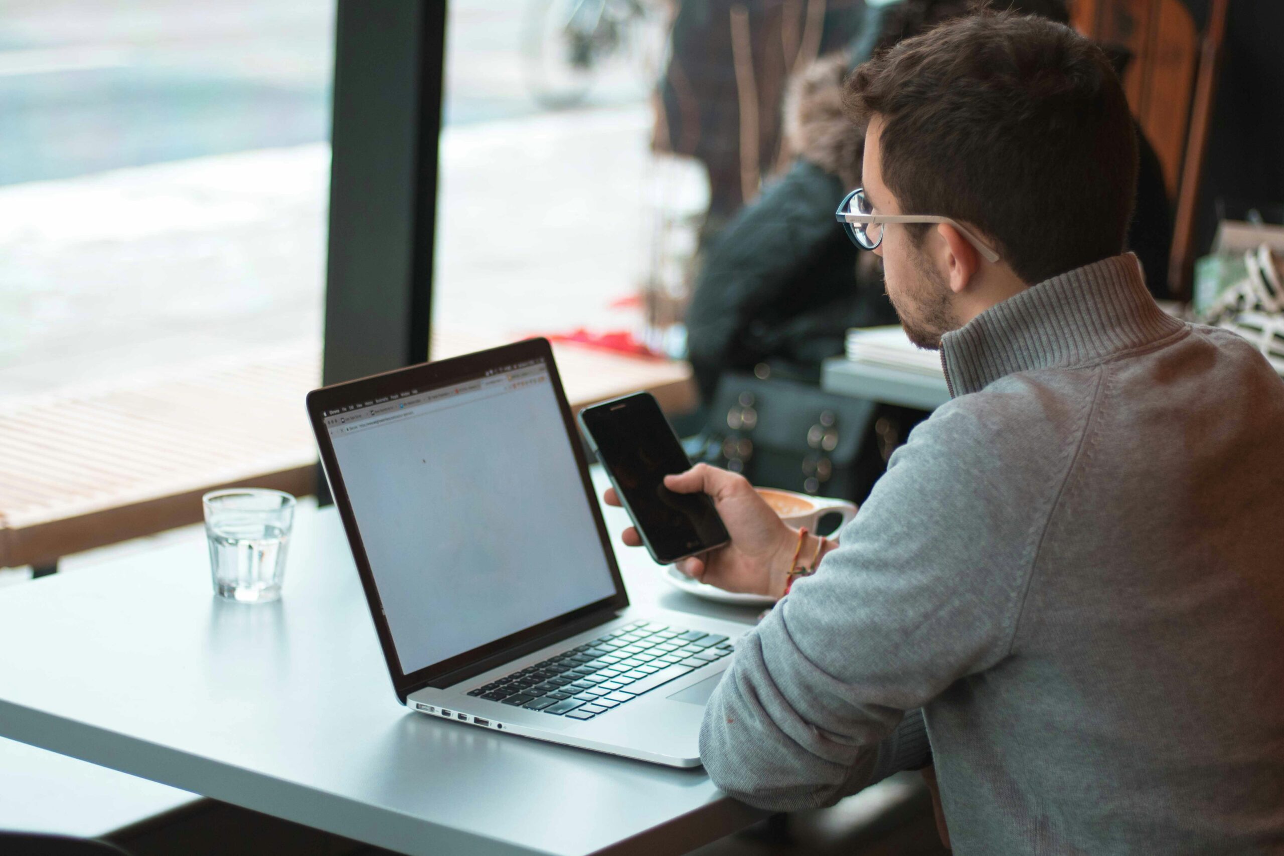Man in glasses working on a laptop while referencing his phone at a café, representing the process of optimizing on-page SEO elements for better e-commerce performance.